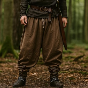 May include: A person wearing brown medieval-style pants with a drawstring waist and matching boots. The outfit includes a brown leather belt with a circular buckle and a long, brown sheath. The background is a forest.