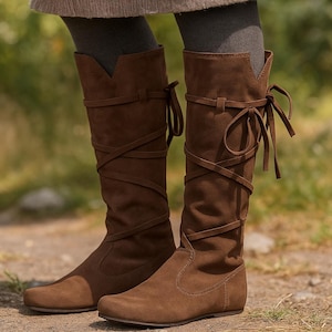 Op de afbeelding: Bruine kniehoge laarzen van suède met kruisende banden en decoratieve strikken. De laarzen hebben een platte zool en een zacht, getextureerd uiterlijk, geschikt voor casual wear. De laarzen worden buiten getoond.