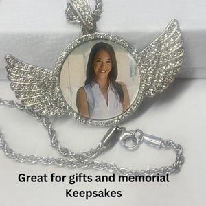 Op de afbeelding: Zilveren ketting met hanger met een ronde fotolijst omringd door heldere strass steentjes. De lijst is bevestigd aan twee vleugels met heldere strass steentjes. De ketting heeft een zilveren touwketting.