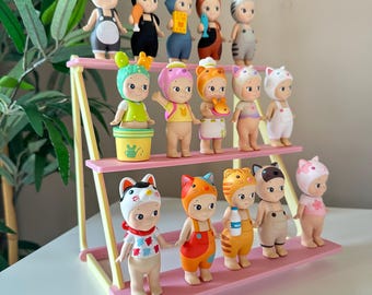 May include: A three-tiered pink display stand holds fifteen small collectible figurines. Each figurine features a unique animal-themed headpiece and various outfits, including overalls, dresses, and accessories like a milk bottle and a watering can.