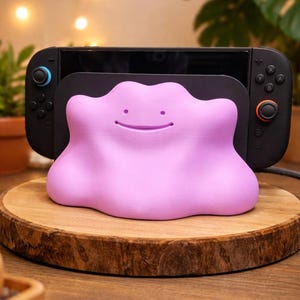 Op de afbeelding: Een lichtpaarse Ditto-vormige Nintendo Switch-dock. De dock staat op een houten schijf, met een zwarte Switch-console op de achtergrond. De dock heeft een lachend gezicht en is ontworpen om de Switch-console vast te houden.