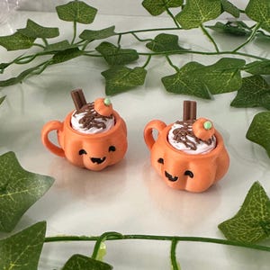 Puede incluir: Dos tazas naranjas con forma de calabaza con caras sonrientes, rellenas de crema batida, salsa de chocolate y una pequeña guarnición de calabaza. Un palito de canela está en la crema batida. Las tazas están decoradas con hojas verdes.