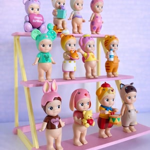 Könnte beinhalten: Eine Sammlung von elf kleinen, stehenden Figuren mit verschiedenen tierischen Kopfbedeckungen und Outfits, präsentiert auf einem dreistufigen, rosa-gelben Holzregal. Die Figuren haben verschiedene Farben, darunter Rosa, Grün, Braun und Orange, wobei eine ein Herz mit der Aufschrift Happy Birthday und eine andere ein T-Shirt mit der Aufschrift LOVE trägt.