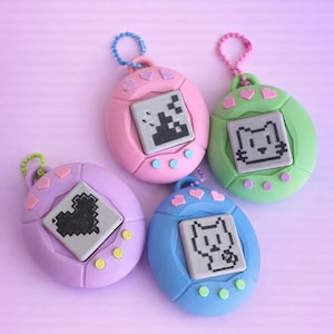 May include: Four brightly coloured digital pet toys in pink, green, blue, and purple. Each toy has a pixelated graphic on a grey screen and three buttons below. The toys have a loop for attaching a keyring.
