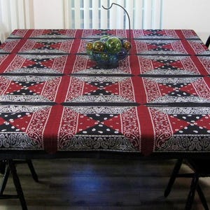 May include: A black and red bandana patterned tablecloth with a white border covers a wooden table with six black chairs. A bowl of decorative glass orbs sits in the center of the table.