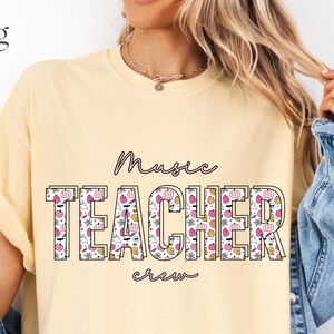 Puede incluir: Una camiseta de manga corta de color amarillo pálido con el texto Music TEACHER crew en letras negras. La palabra TEACHER está rellena con un patrón repetitivo de pequeñas calabazas rosas y naranjas, fantasmas blancos y murciélagos negros.