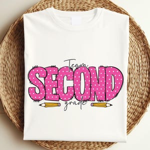 Puede incluir: Camiseta blanca con un diseño de lunares rosa y blanco que dice "Team Second Grade" con dos lápices amarillos debajo.