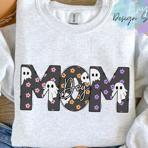 Pode incluir: Uma camisa de frio cinza com a palavra "MOM" em letras pretas com um padr&atilde;o de fantasmas e flores. O "O" de MOM &eacute; substitu&iacute;do pela palavra "Dog" em letras laranja e pretas.