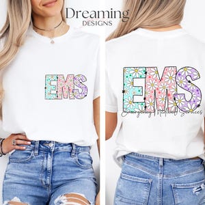 Puede incluir: Camiseta blanca con las letras "EMS" en un diseño floral. Las letras están delineadas en negro y rellenas con una variedad de colores pastel y flores de margarita blancas. El texto "Emergency Medical Services" está escrito debajo de las letras.