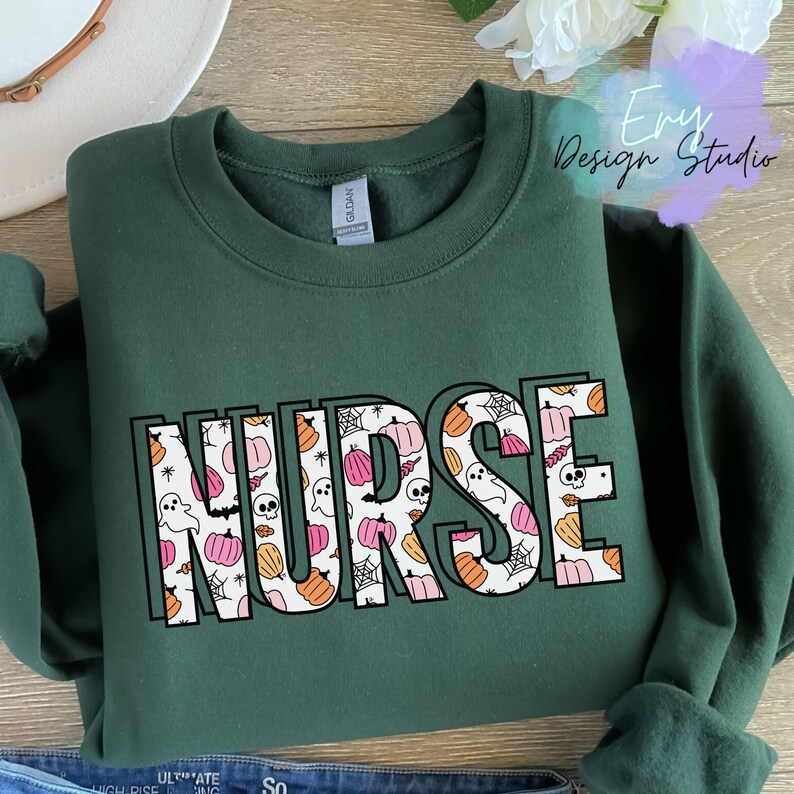 Puede incluir: Una sudadera verde oscuro con la palabra "NURSE" en letras grandes y contorneadas. Las letras est&aacute;n rellenas con un patr&oacute;n de calabazas, fantasmas, telara&ntilde;as y calaveras en naranja, rosa y blanco, ideal para Halloween.