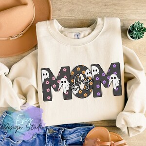 Pode incluir: Uma camisola de cor creme com a palavra "MOM" em letras pretas delineadas. As letras s&atilde;o decoradas com um padr&atilde;o de flores roxas e cor-de-rosa e fantasmas brancos. O "O" de MOM &eacute; substitu&iacute;do por um c&iacute;rculo preto com bolinhas laranja e pretas e a palavra "Dog" escrita em it&aacute;lico preto.