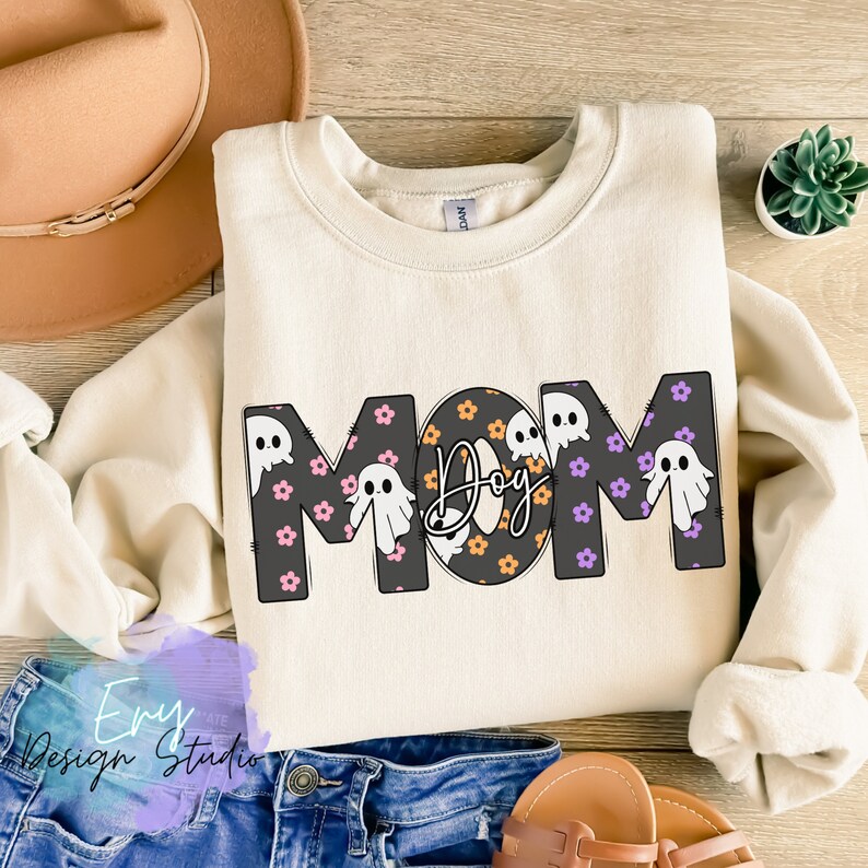 Pode incluir: Uma camisola de cor creme com a palavra "MOM" em letras pretas delineadas. As letras s&atilde;o decoradas com um padr&atilde;o de flores roxas e cor-de-rosa e fantasmas brancos. O "O" de MOM &eacute; substitu&iacute;do por um c&iacute;rculo preto com bolinhas laranja e pretas e a palavra "Dog" escrita em it&aacute;lico preto.