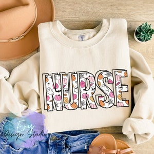 Puede incluir: Sudadera color crema con la palabra "NURSE" en contorno negro, rellena de patrones de Halloween. Los patrones incluyen calabazas, fantasmas y telara&ntilde;as. Se ve un sombrero beige y unos vaqueros azules.