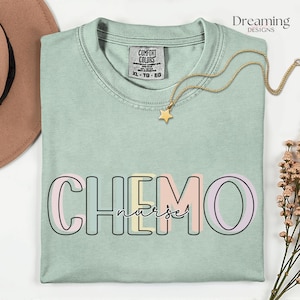 May include: A pale green Comfort Colors t-shirt featuring the word "CHEMO" in pastel colours, with "nurse" below. A gold star pendant necklace and a brown hat are also visible in the image.
