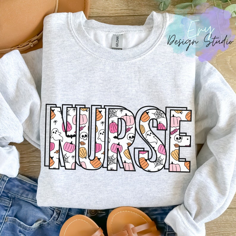Puede incluir: Una sudadera gris claro con la palabra "NURSE" en letras grandes y delineadas. Las letras están rellenas con un patrón de calabazas, fantasmas, telarañas y calaveras en naranja, rosa y blanco, ideal para Halloween.