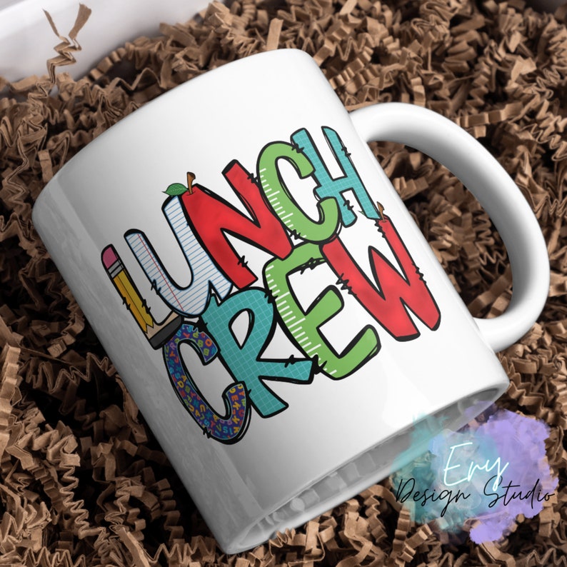 Może przedstawiać: Biały kubek ceramiczny z kolorowym wzorem graficznym, na kt&oacute;rym widnieje napis "Lunch Crew". Wz&oacute;r zawiera czerwone jabłko, oł&oacute;wek i książkę.
