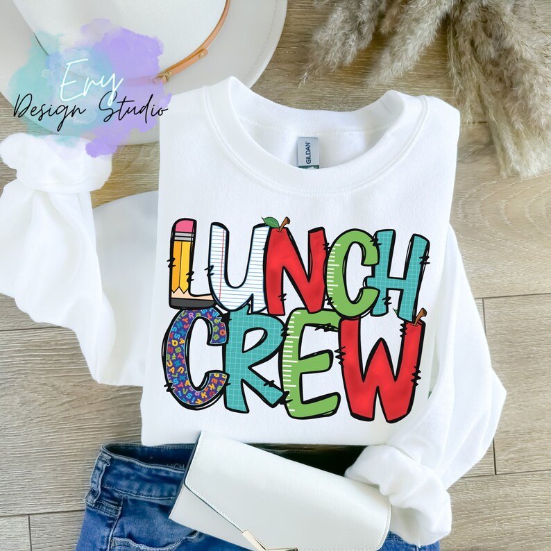 Może przedstawiać: Biała bluza z napisem "Lunch Crew" w kolorowych, zabawnych literach. Litery są ozdobione wzorami w szkolnym stylu, takimi jak oł&oacute;wki, jabłka i papier w kratkę.