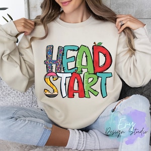 May include: A beige sweatshirt with the words "HEAD START" in colorful, playful letters. The letters are decorated with school-themed patterns like pencils, rulers, and alphabet blocks.