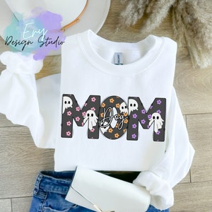 Pode incluir: Camisola branca com a palavra "MOM" em letras pretas delineadas. O "O" &eacute; substitu&iacute;do por um c&iacute;rculo preto e laranja com a palavra "Dog" dentro. As letras s&atilde;o decoradas com flores cor-de-rosa e roxas e fantasmas brancos.