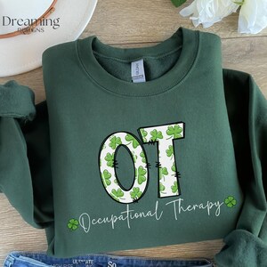 以下が含まれることがあります： 白と緑のクローバー模様の文字「OT」が入った濃い緑色のスウェットシャツ。「Occupational Therapy」の文字が筆記体で下に書かれています。言葉の右側にはクローバーがあります。