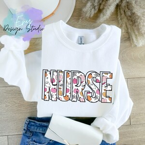 Puede incluir: Sudadera blanca de cuello redondo con la palabra "NURSE" en un contorno negro. Las letras est&aacute;n rellenas con un patr&oacute;n de calabazas, fantasmas y telara&ntilde;as en naranja, rosa y blanco. La sudadera est&aacute; doblada sobre unos vaqueros azules.