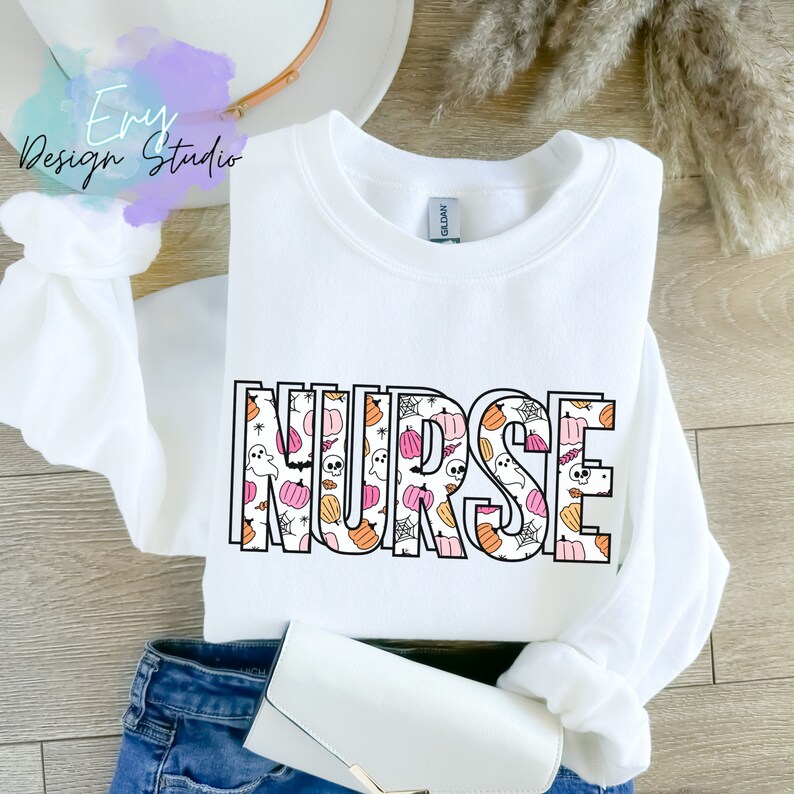 Puede incluir: Sudadera blanca de cuello redondo con la palabra "NURSE" en un contorno negro. Las letras est&aacute;n rellenas con un patr&oacute;n de calabazas, fantasmas y telara&ntilde;as en naranja, rosa y blanco. La sudadera est&aacute; doblada sobre unos vaqueros azules.