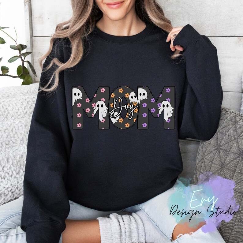Pode incluir: Camisola de moletom preta com a palavra "MOM" em letras grandes, com um padr&atilde;o de fantasmas e a palavra "Boo" no "O".