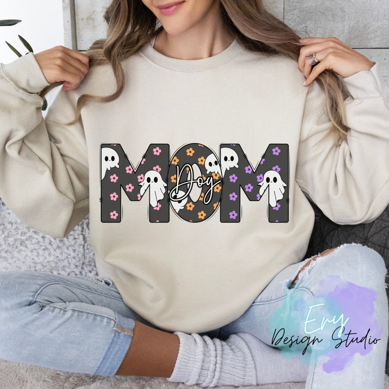 Pode incluir: Uma camisa de moletom bege com a palavra "MOM" em letras pretas. As letras s&atilde;o decoradas com pequenas flores roxas e rosas e silhuetas de fantasmas brancas. O "O" de MOM &eacute; um c&iacute;rculo preto com flores laranja e brancas e a palavra "Boo" escrita em branco.