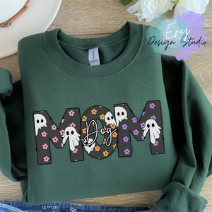 Pode incluir: Camisola verde com a palavra "MOM" em letras grandes. As letras s&atilde;o decoradas com pequenos fantasmas e flores. A palavra "Dog" est&aacute; no centro das letras.