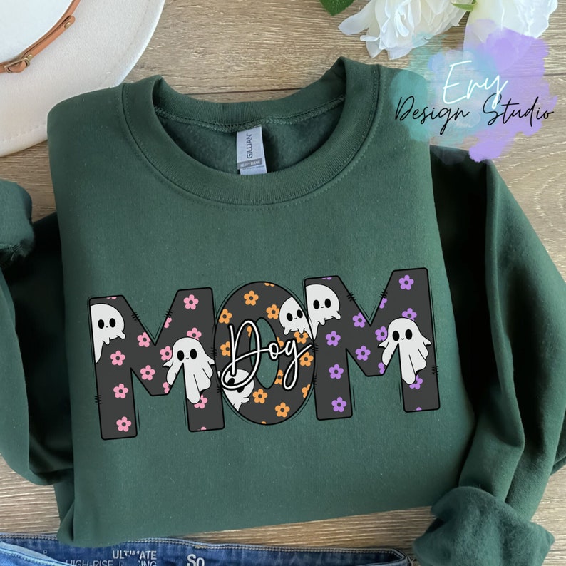 Pode incluir: Camisola verde com a palavra "MOM" em letras grandes. As letras s&atilde;o decoradas com pequenos fantasmas e flores. A palavra "Dog" est&aacute; no centro das letras.