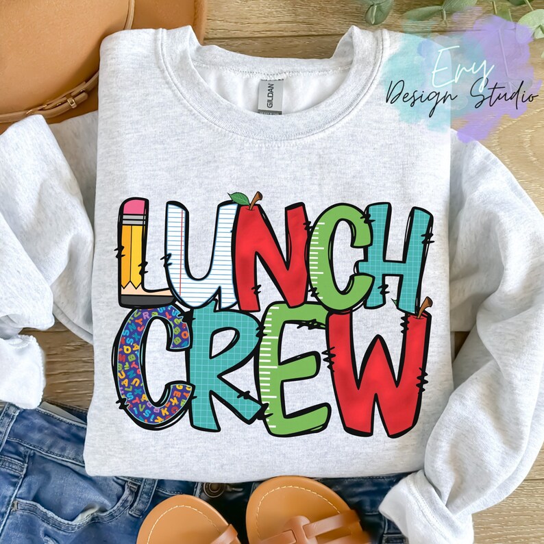 Może przedstawiać: Szara bluza z napisem "Lunch Crew" w kolorowych, kresk&oacute;wkowych literach. Litery są ozdobione wzorami w szkolnym stylu, takimi jak oł&oacute;wki, jabłka i klocki alfabetu.