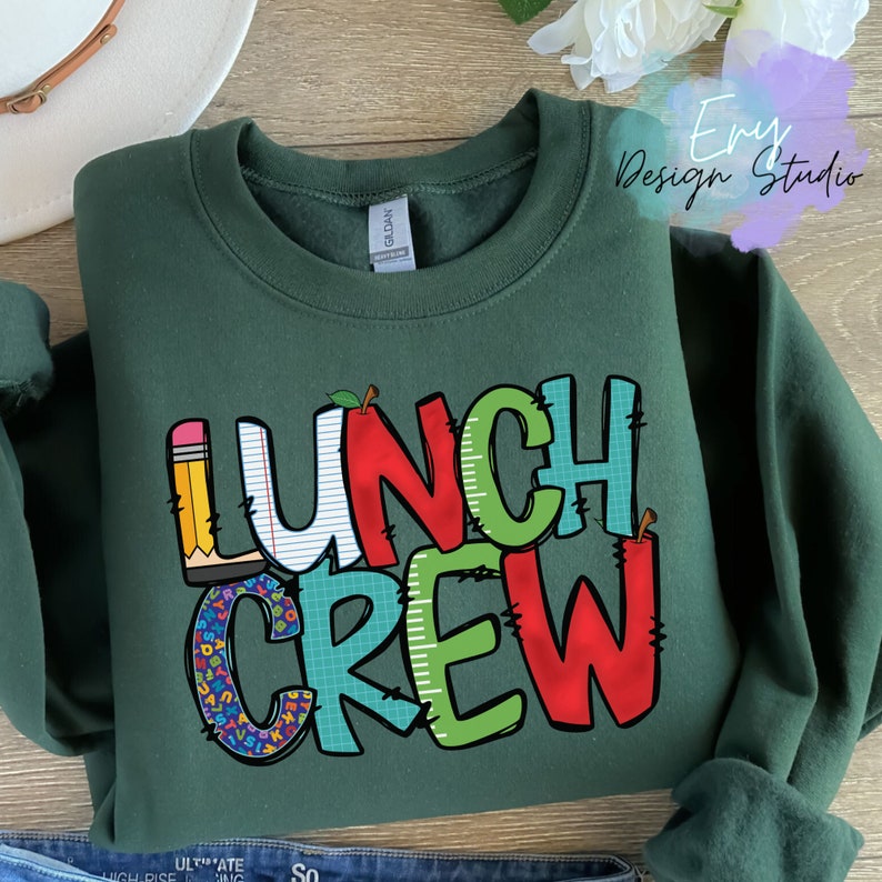 Może przedstawiać: Zielony sweter z napisem "Lunch Crew" w kolorowych literach. Litery są ozdobione przyborami szkolnymi, takimi jak oł&oacute;wki, papier i jabłka.
