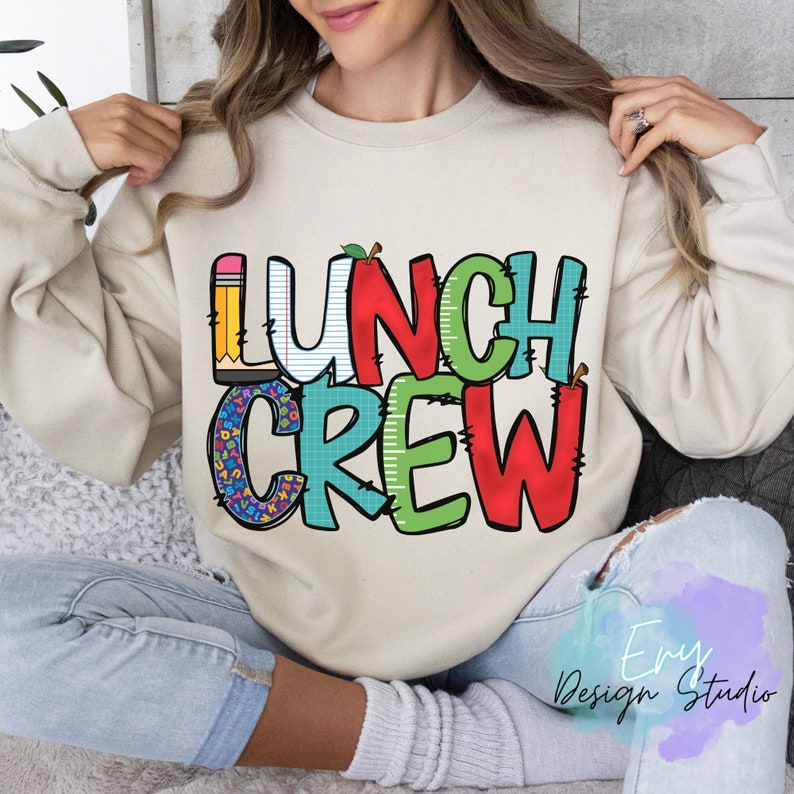 Może przedstawiać: Beżowa bluza z napisem "LUNCH CREW" w kolorowych, zabawnych literach. Litery są ozdobione oł&oacute;wkiem, jabłkiem i kratką.