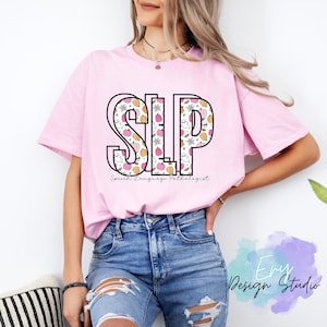 Può includere: Maglietta rosa con un contorno nero delle lettere "SLP" riempite con un motivo di zucche arancioni e rosa, fantasmi e ragnatele. Il testo "Speech Language Pathologist" è sotto le lettere.