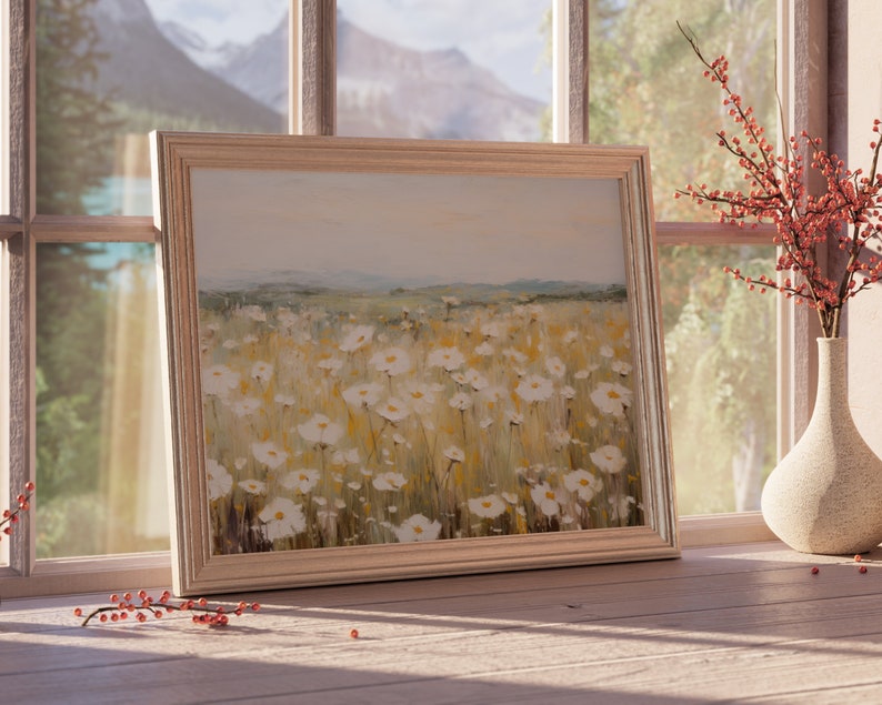 May include: A framed painting of a field of white wildflowers with a blurred background of a mountain range and a blue sky. The frame is light wood and the painting is hanging on a wall.