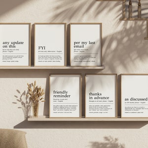 May include: Six minimalist black and white printable wall art prints with definitions of common office phrases. The prints are framed in light wood frames and displayed on a shelf against a light beige wall.
