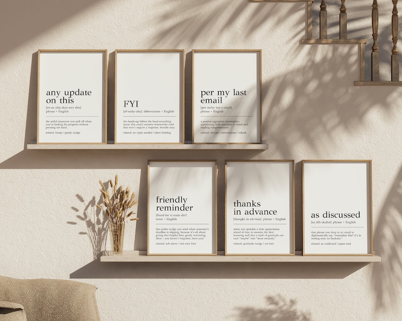 May include: Six minimalist black and white printable wall art prints with definitions of common office phrases. The prints are framed in light wood frames and displayed on a shelf against a light beige wall.