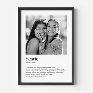 May include: Black and white framed print featuring a photograph of two smiling women embracing. The word "bestie" is displayed below the photo, along with a definition. The frame is black and the background is white.