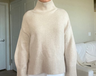 May include: A cream-colored turtleneck sweater with long sleeves and a ribbed hem. The sweater is made of a soft, knit fabric.