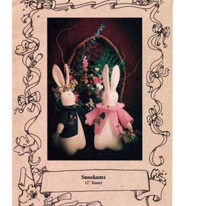 May include: Two plush bunny dolls, one in a black sweater and the other in a pink jacket, stand before a floral arrangement. The text "Snookums 12" Bunny" is displayed below the dolls. The image is framed with floral and ribbon designs.
