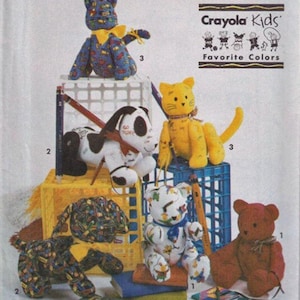 May include: A collection of handmade fabric stuffed animals, including a blue cat, a yellow cat, a black and white dog, a brown bear, and a white bear. The toys are displayed on and around blue and yellow crates. The image includes the text "Crayola Kids Favorite Colors."