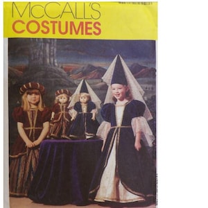 Puede incluir: Patrón de disfraces McCall's con disfraces medievales para niños. La imagen muestra a una niña y muñecas con vestidos azul oscuro y blanco con adornos dorados, y una niña con un vestido burdeos. El fondo presenta un castillo y montañas.