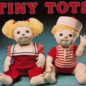 May include: Two cloth dolls dressed in sailor outfits. The girl doll has blonde hair with pigtails and is wearing a red and white striped shirt with a white sailor collar and red overalls. The boy doll has blonde hair and is wearing a red and white striped shirt with a white sailor hat. The dolls are sitting on a blue surface. The text "TINY TOTS" is written in red letters above the dolls.