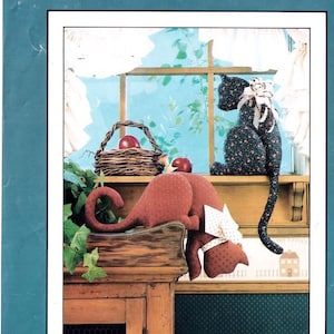 Op de afbeelding: Twee stoffen katten, een rode en een zwarte met bloempatronen, worden tentoongesteld op een houten plank. Een mand met rode appels staat in de buurt. De scène speelt zich af voor een raam met uitzicht op een blauwe lucht.