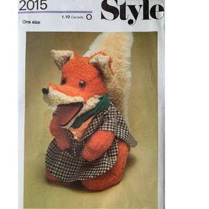 以下が含まれることがあります： 茶色と白のチェック柄のコートと緑色のスカーフをつけた、ぬいぐるみ製のキツネの人形。キツネはオレンジ色の大きな体、白い尻尾、茶色の鼻をしています。