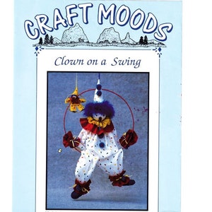 Könnte beinhalten: Ein Bastelmuster für eine Clown-Puppe auf einer Schaukel. Der Clown trägt ein weißes und rotes Kostüm mit blauen und gelben Akzenten. Das Muster trägt den Titel "Clown on a Swing" und stammt von Craft Moods.