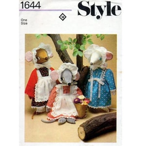 Può includere: Un modello di cucito per tre bambole topo. Il modello si chiama "Style" e il numero è 1644. Le bambole indossano abiti e grembiuli. Una bambola indossa un vestito rosso con un grembiule bianco, un'altra indossa un vestito blu con un grembiule bianco e la terza indossa un vestito bianco con un grembiule rosso. Le bambole sono in piedi su un tronco.