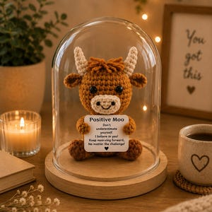 May include: A brown crocheted highland cow figurine with cream-coloured horns and a white muzzle, enclosed in a glass dome on a wooden base. The cow holds a white square sign that reads: Positive Moo, Don't underestimate yourself. I believe in you! Keep moo-ving forward, No matter the challenge!