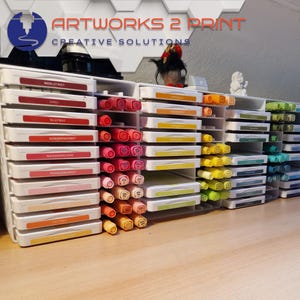 Op de afbeelding: Een verzameling inktkussens en stiften in diverse kleuren, netjes geordend in witte plastic opbergeenheden. De inktkussens hebben labels met Duitse kleurnamen. De afbeelding toont ook de tekst "ARTWORKS 2 PRINT CREATIVE SOLUTIONS".