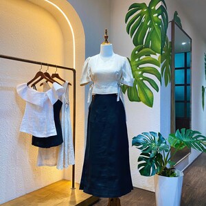 Puede incluir: Una blusa blanca de lino con mangas cortas y una falda negra de lino. La blusa tiene un detalle de lazo en la cintura. La falda tiene una cintura alta y un corte holgado. El atuendo se muestra en un maniqu&iacute; en un entorno de tienda.